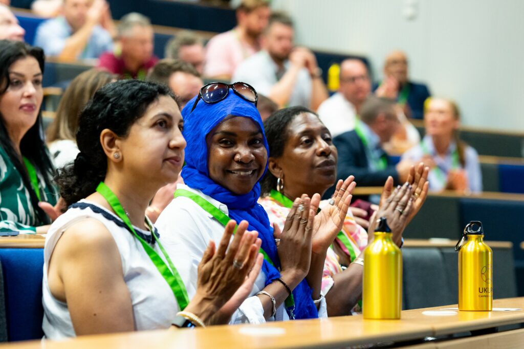 A crowd of delegates at a conference clapping. The woman in the middle is smiling at the camera.